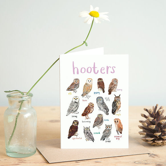 Card with owl illustrations and text 'hooters' on a wooden surface with a vase and pine cone.