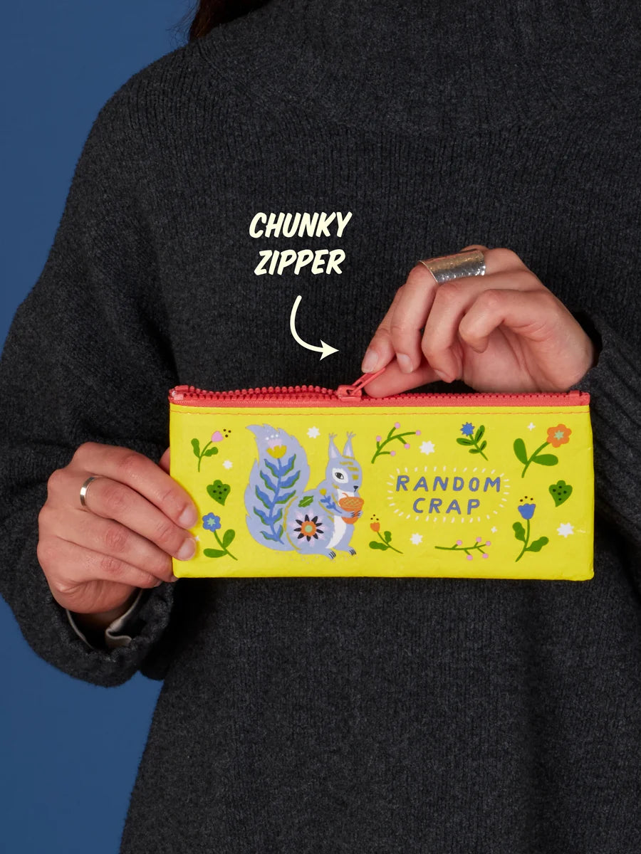 A person wearing a dark gray sweater holds a bright yellow pencil case with a whimsical squirrel illustration and the words 'RANDOM CRAP.' White text on the image points to the zipper, which is labeled 'CHUNKY ZIPPER.