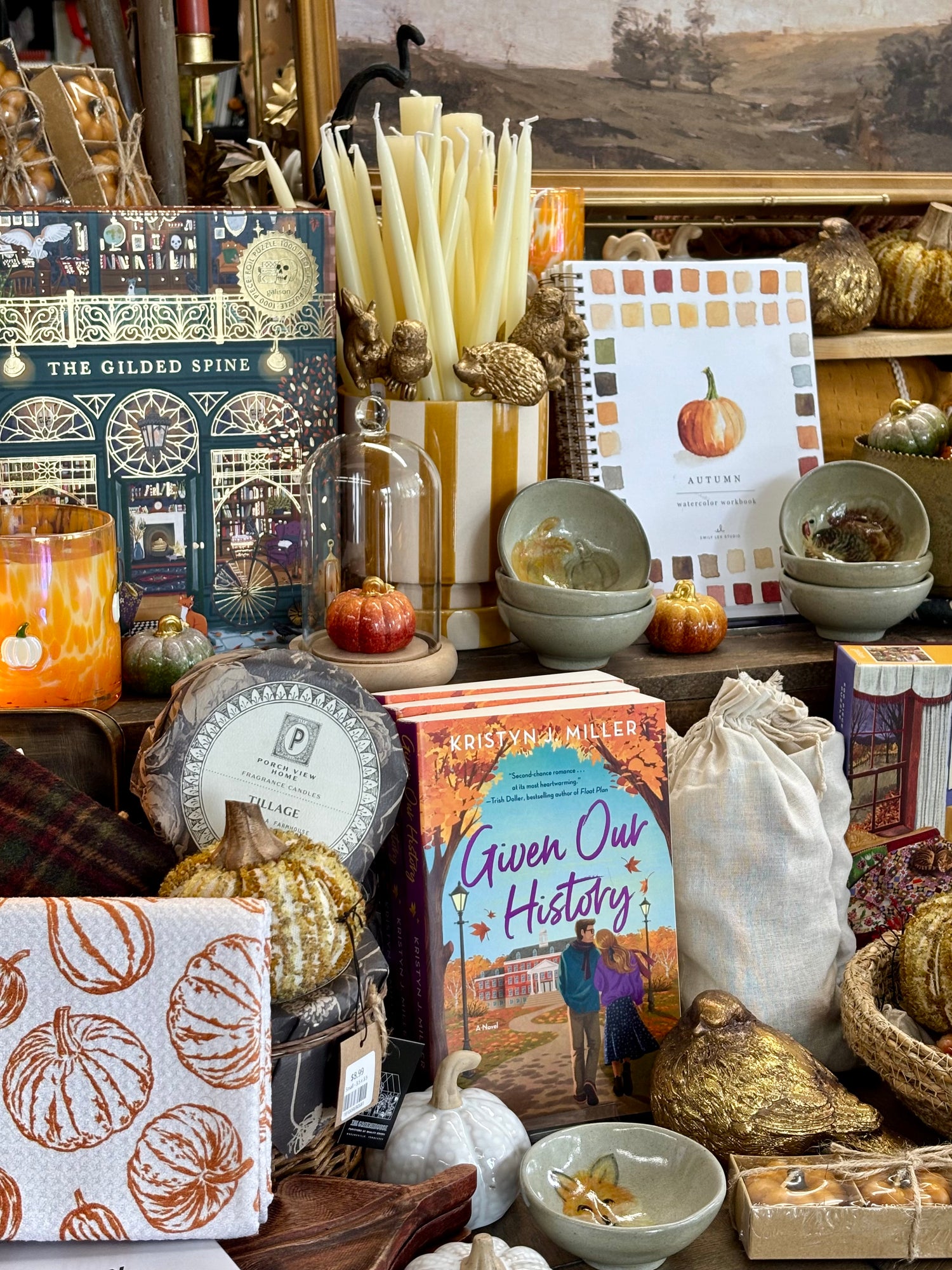 A styled autumn-themed gift table display featuring a book titled 'Given Our History' by Kristin Millar, a candle tin labeled 'The Gilded Spine', ceramic bowls, pumpkins, candles, and other seasonal home decor items.