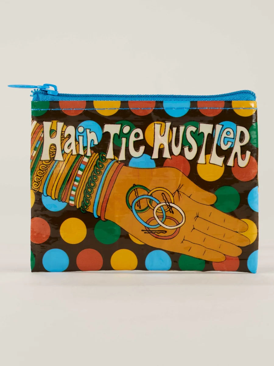 A zippered pouch featuring a colorful graphic print with the text 'Hair Tie Hustler' and an illustration of a hand wearing colorful hair ties and rings, set against a light gray background.