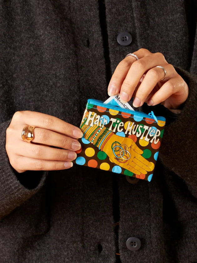 A person wearing a dark charcoal knit cardigan holds a small colorful coin purse labeled 'HATE THE MISTLE' against a solid purple background.