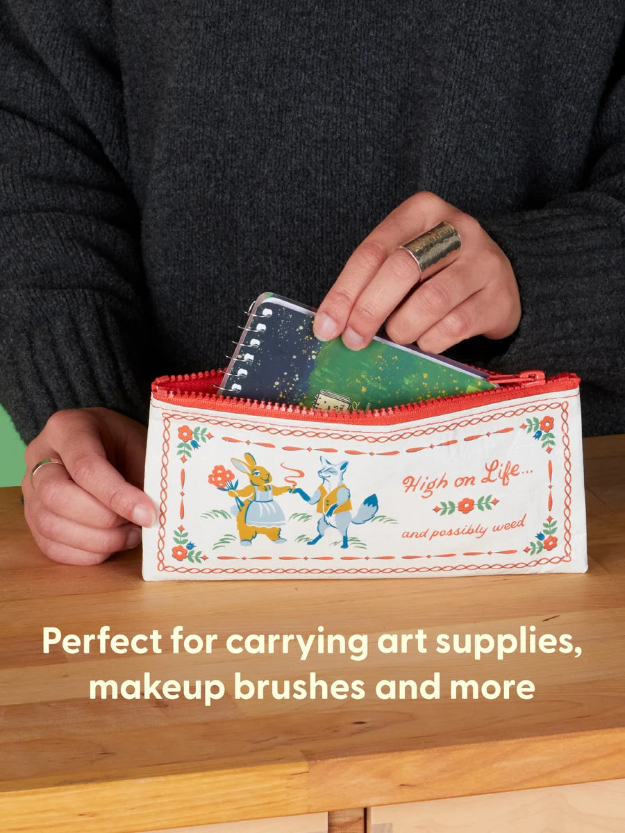 A person holds a small decorative zip pouch on a wooden table, inserting a notebook into the pouch. The pouch is white with a red zipper, embroidered-style floral border, and cartoon rabbit illustrations with the text 'High on Life.