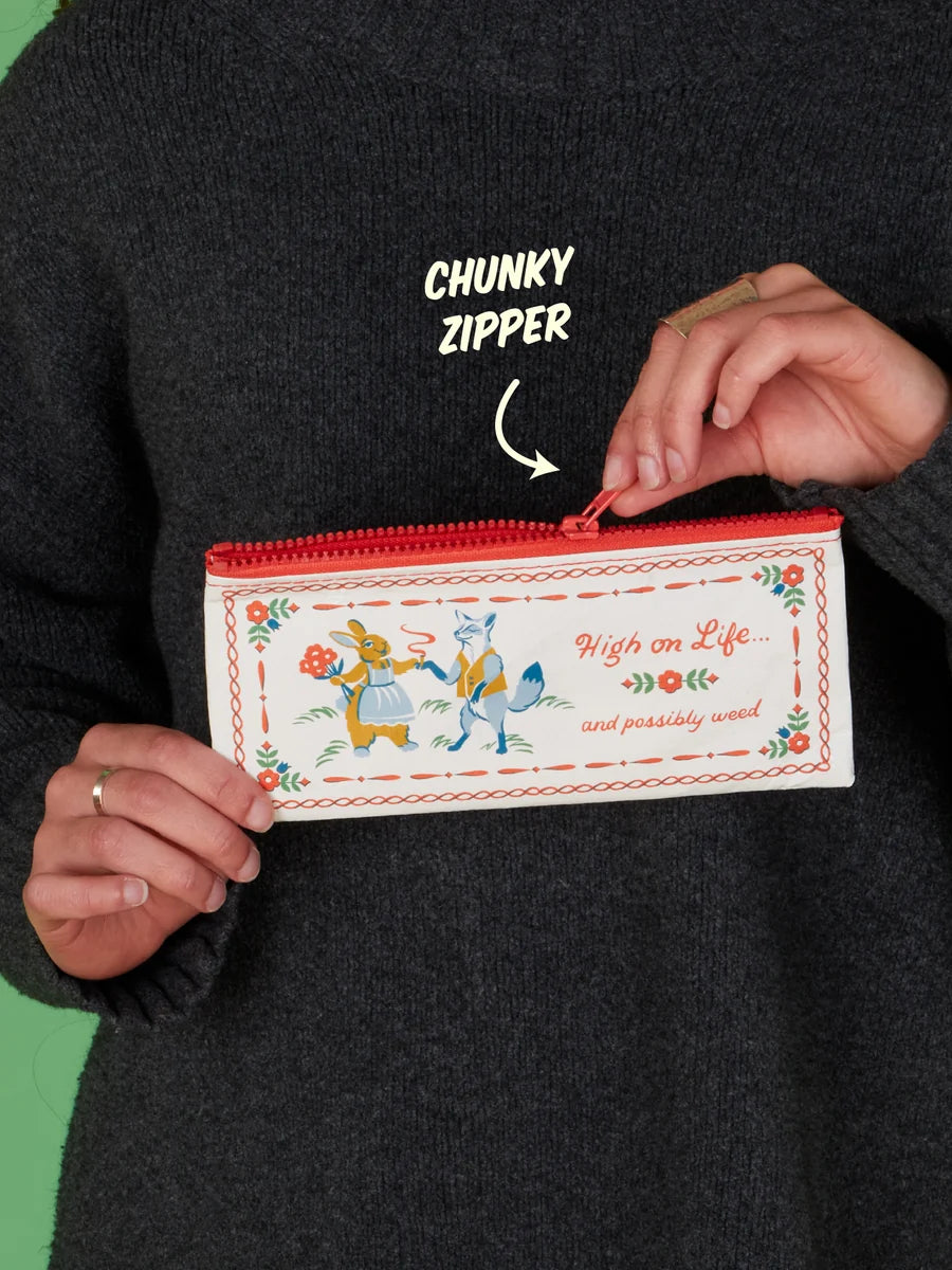 A person in a dark gray sweater holds a rectangular zipper pouch with a red zipper and a white front decorated with colorful cartoon animals and the words 'High on Life and possibly dead.' White text above reads 'CHUNKY ZIPPER' with an arrow pointing to the zipper.