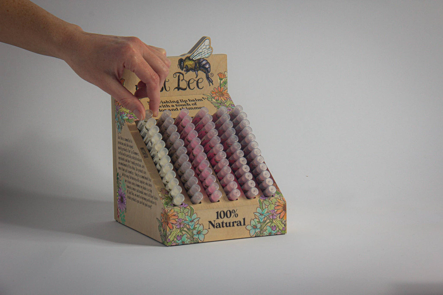 Hand reaching into a box of natural lip shimmers with a visible brand name on a gray background