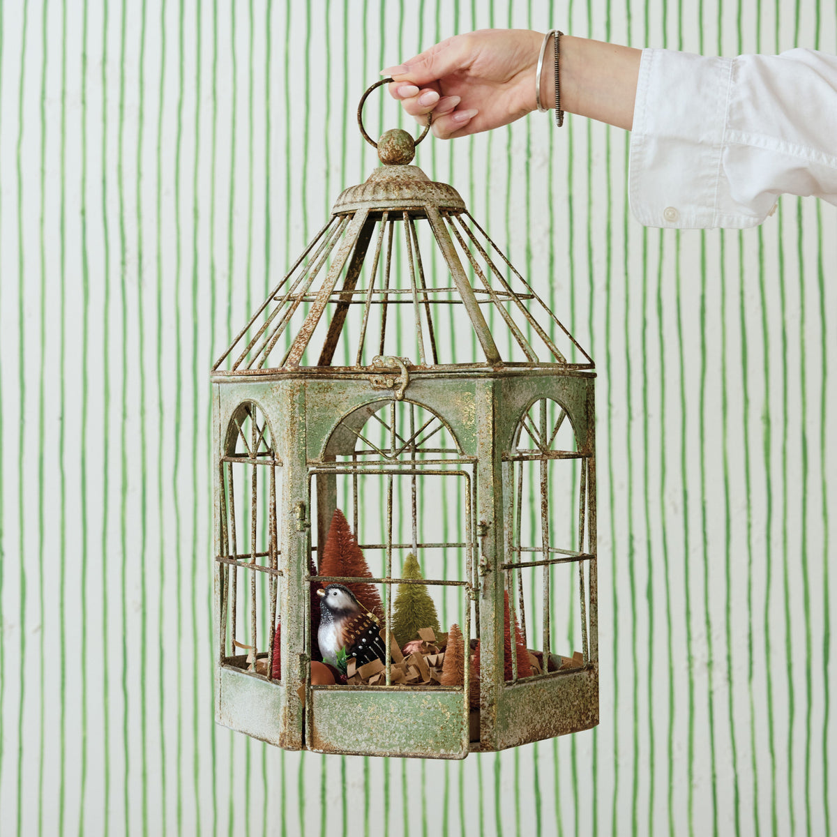 A hand holds a distressed green metal birdcage decoration with a miniature bird and Christmas tree inside, set against a green and white vertically striped wallpaper background.