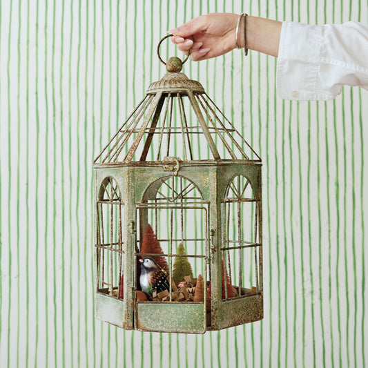A hand holds a distressed green metal birdcage decoration with a miniature bird and Christmas tree inside, set against a green and white vertically striped wallpaper background.