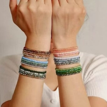 A close-up of a person's forearms and hands wearing multiple beaded bracelets. The bracelets come in several colors including brown, blue, green, pink, and silver, stacked on both wrists against a light neutral background.
