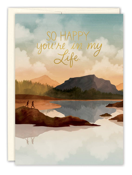Greeting card with a scenic illustration of a lake and mountains, featuring the text 'So happy you're in my life' in gold.