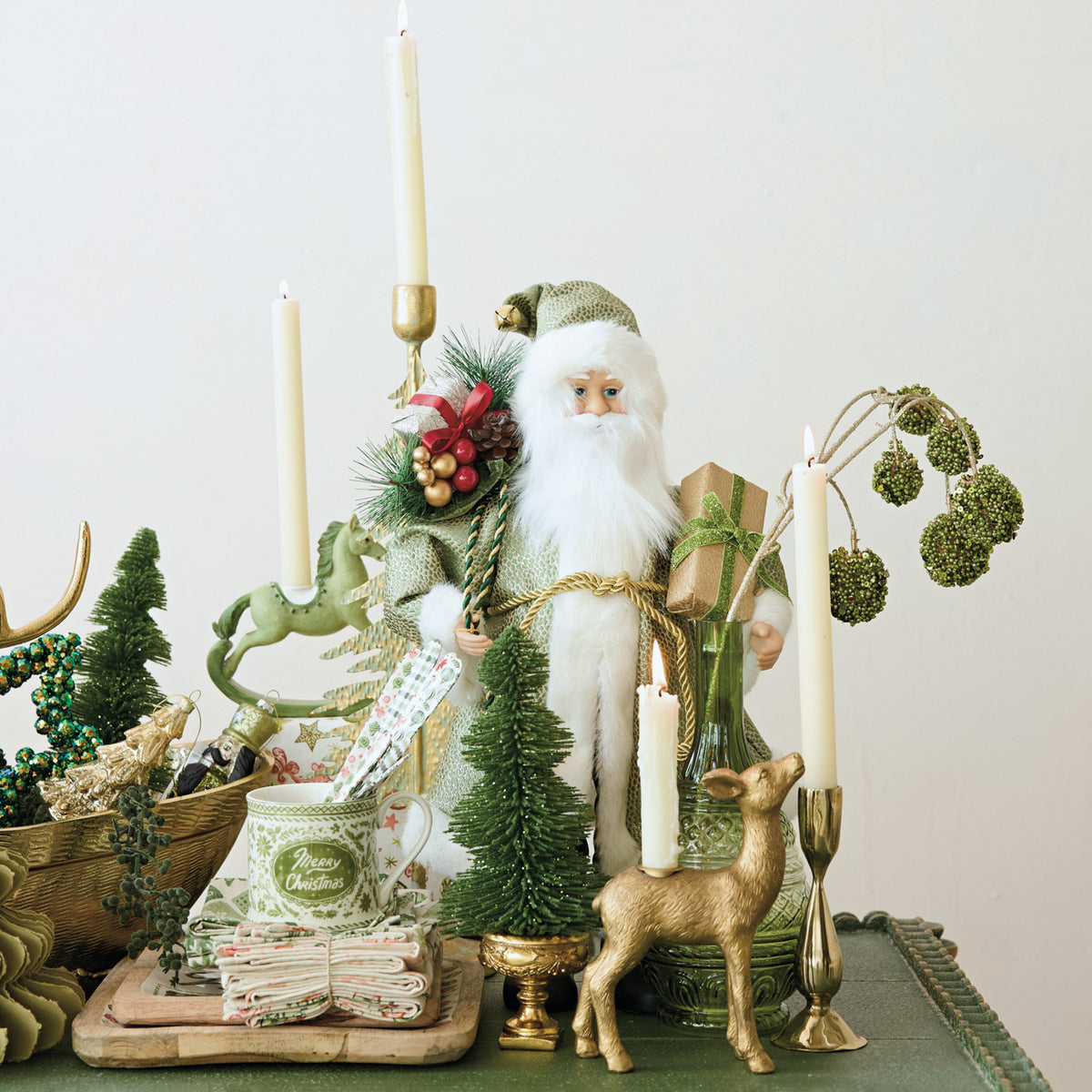 Decorative Christmas scene with a Santa figure, candles, and greenery on a table.