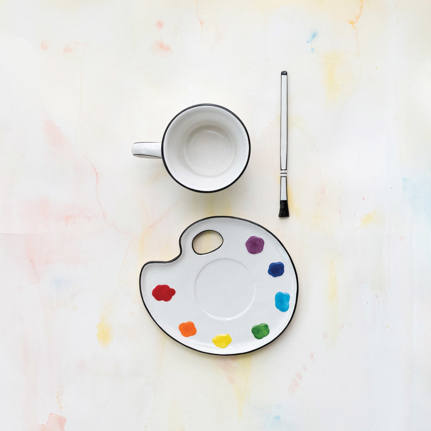Empty mug, paint palette with colorful dots, and paintbrush on a light background