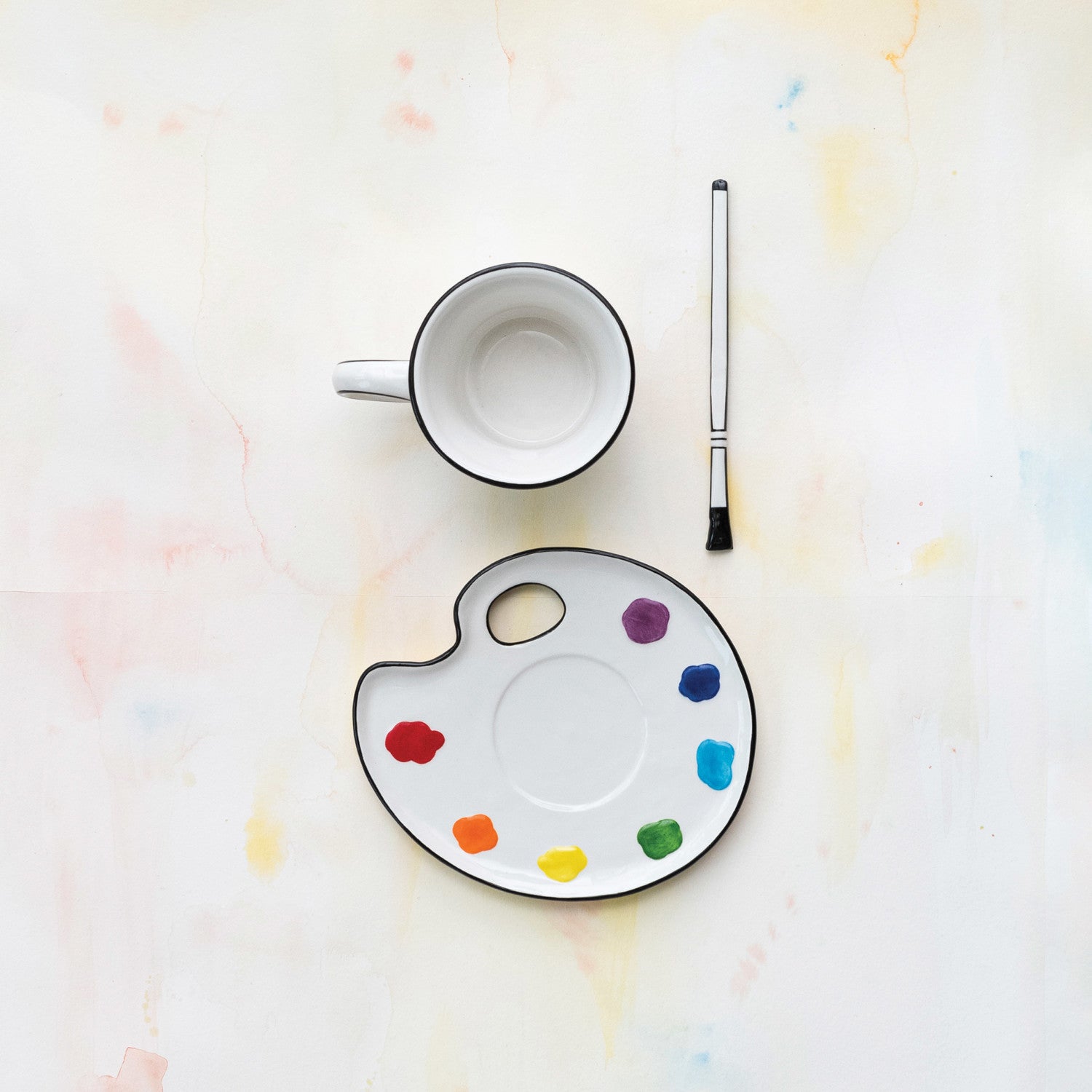 Empty mug, paint palette with colorful dots, and paintbrush on a light background