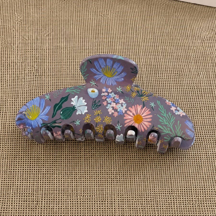 A purple hair claw clip decorated with a colorful hand-painted floral pattern, resting on a textured beige woven mat.