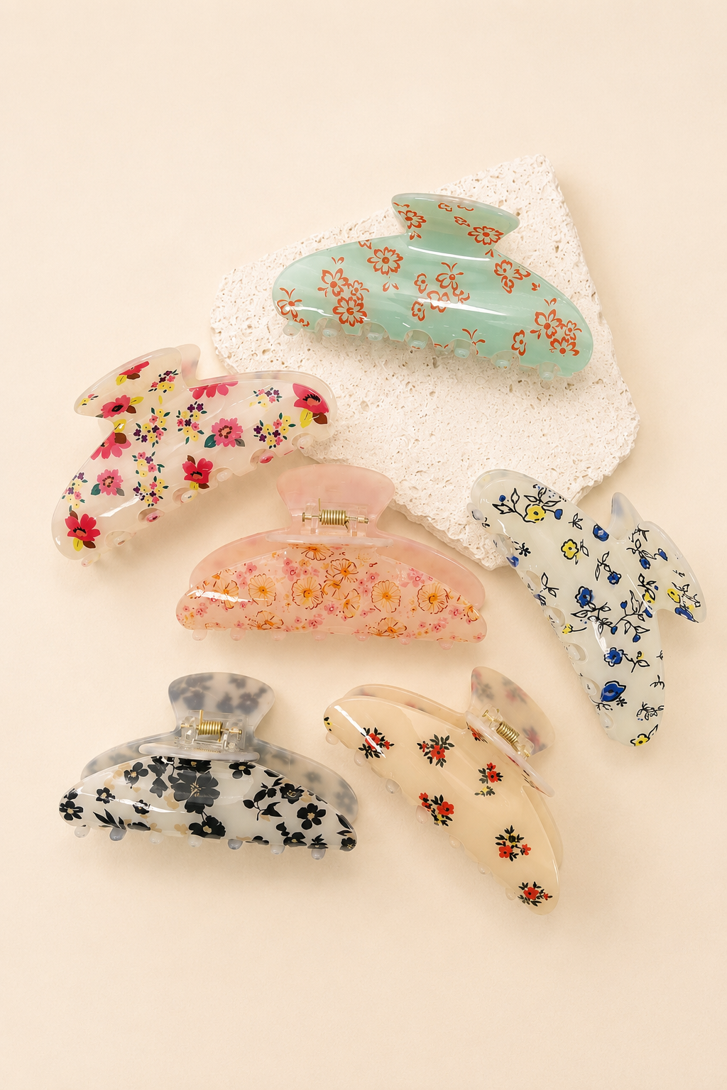 Six floral hair claw clips in various colors and patterns, arranged on a light beige background with a white textured paper accent.