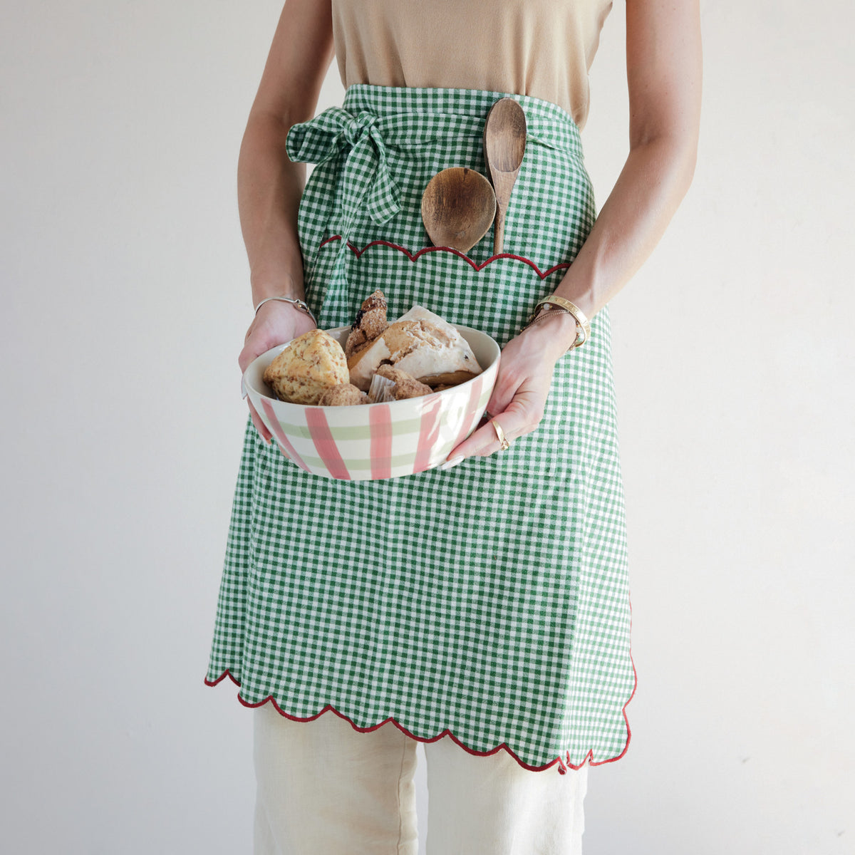 Green gingham cotton scalloped half apron with red stitched trim and front pocket worn while holding a bowl of baked goods.