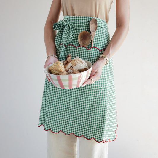 Green gingham cotton scalloped half apron with red stitched trim and front pocket worn while holding a bowl of baked goods.