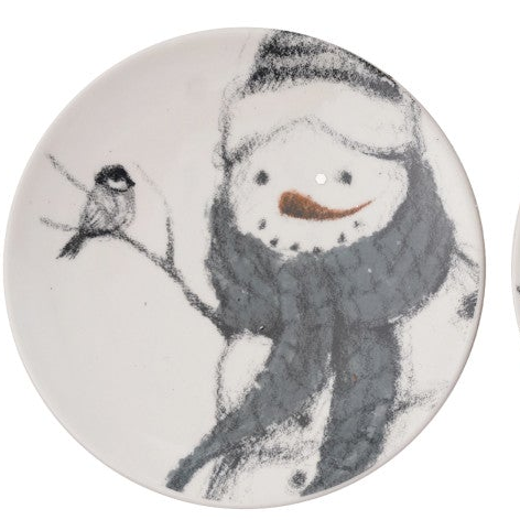 A round ceramic plate featuring a whimsical hand-painted style illustration of a snowman wearing a striped beanie and a dark gray scarf, with a small black bird perched on a branch to the left. The plate has a warm off-white glaze background.
