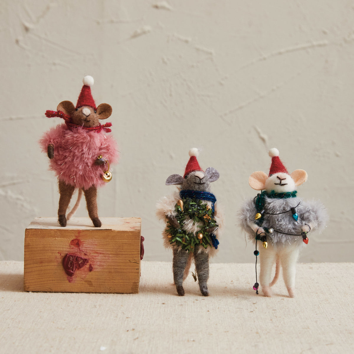 Three wool felt Christmas mice wearing Santa hats and fluffy faux fur sweaters decorated with colorful garland, bells, beads, and miniature holiday lights.